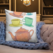 Load image into Gallery viewer, A white pillow on a sofa with a blue knitted blanket. The white pillow features the artwork on a white background with four teacups on saucers and one large teapot. The teacups are in muted colors of orange, blue, yellow and green and the teapot is a muted orange. On the teacups, saucers and teapot there is a light flower detail pattern.
