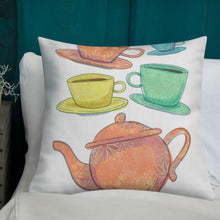 Load image into Gallery viewer, A white pillow with illustrations leading on white bedding with a side table off to the side. The white pillow features the artwork on a white background with four teacups on saucers and one large teapot. The teacups are in muted colors of orange, blue, yellow and green and the teapot is a muted orange. On the teacups, saucers and teapot there is a light flower detail pattern.