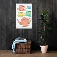 Load image into Gallery viewer, A canvas is hanging on a dark wood panelled wall with a basket with a blanket and a potted plant on the floor. The artwork is on a white background with four teacups on saucers and one large teapot. The teacups are in muted colors of orange, blue, yellow and green and the teapot is a muted orange. On the teacups, saucers and teapot there is a light flower detail pattern.
