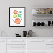 Load image into Gallery viewer, A black frame is hanging on a stucco kitchen wall above a kitchen counter and next to some shelves. The white shelves have clear containers of pasta and the bottom shelf has tea mugs. The counter has a tea pot and plates on it. The frame has artwork on a white background with four teacups on saucers and one large teapot. The teacups are in muted colors of orange, blue, yellow and green and the teapot is a muted orange. On the teacups, saucers and teapot there is a light flower detail pattern.