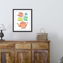 Load image into Gallery viewer, A black frame is hanging above a brown credenza with a lamp and basket on top of it. The frame contains artwork on a white background with four teacups on saucers and one large teapot. The teacups are in muted colors of orange, blue, yellow and green and the teapot is a muted orange. On the teacups, saucers and teapot there is a light flower detail pattern.