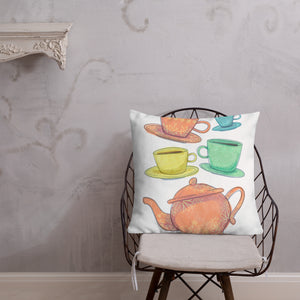 A pillow on a chair against a grey wall. The white pillow features the artwork on a white background with four teacups on saucers and one large teapot. The teacups are in muted colors of orange, blue, yellow and green and the teapot is a muted orange. On the teacups, saucers and teapot there is a light flower detail pattern.