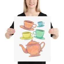 Load image into Gallery viewer, A woman is holding a paper art print in her hands. The artwork is on a white background with four teacups on saucers and one large teapot. The teacups are in muted colors of orange, blue, yellow and green and the teapot is a muted orange. On the teacups, saucers and teapot there is a light flower detail pattern.