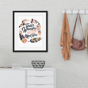 Artwork featured on a wall in a black frame by a shelving unit. The artwork is on white paper and features hand drawn lettering with the words "Tender Warriors Are Being Made Here" The words are in pink, navy and dark mustard yellow. 