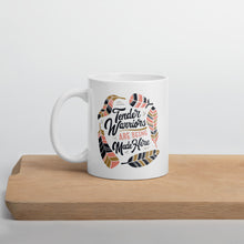 Load image into Gallery viewer, A white mug sitting on a piece of wood. The white mug features hand drawn lettering with the words &quot;Tender Warriors Are Being Made Here&quot; The words are in pink, navy and dark mustard yellow. 
