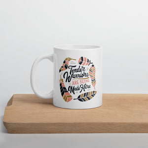 A white mug sitting on a piece of wood. The white mug features hand drawn lettering with the words "Tender Warriors Are Being Made Here" The words are in pink, navy and dark mustard yellow. 