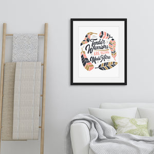 Framed artwork in a black frame on a wall above a sofa featuring a white paper print with hand drawn lettering and illustrations with the words "Tender Warriors Are Being Made Here" The words are in pink, navy and dark mustard yellow. 