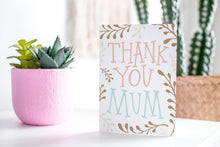 Load image into Gallery viewer, A greeting card featured standing up on a white tabletop with a pink plant pot in the background and some succulents in the pot. There’s a woven basket in the background with a cactus inside. The card features illustrated plant leaves around the words “Thank you Mum.”