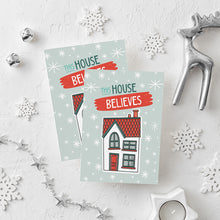 Load image into Gallery viewer, Two Christmas cards laying on a white background with white and silver Christmas decorations on the table. The Christmas card has a light blue background with stars. The words &quot;this house believes&quot; is featured above an illustrated house. The words and house are in white, green and light blue.