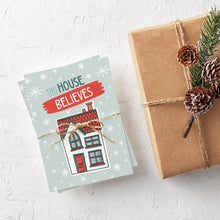 Load image into Gallery viewer, A stack of Christmas cards with brown string wrapped around them. A brown craft paper gift is off to the side. The Christmas card has a light blue background with stars. The words &quot;this house believes&quot; is featured above an illustrated house. The words and house are in white, green and light blue.