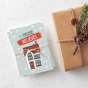 A stack of Christmas cards with brown string wrapped around them. A brown craft paper gift is off to the side. The Christmas card has a light blue background with stars. The words "this house believes" is featured above an illustrated house. The words and house are in white, green and light blue.