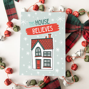 A Christmas card featured on top of some red and white Christmas decorations. The Christmas card has a light blue background with stars. The words "this house believes" is featured above an illustrated house. The words and house are in white, green and light blue.
