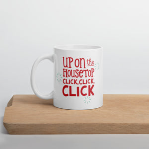 A white mug sitting on a light wood cutting board. The design features the words "Up on the housetop, click, click, click" in red with small blue stars around it.