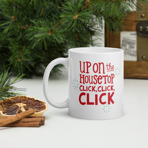 A white mug with a pine tree in the background. The design is in red with the words "Up on the housetop click, click, click" with small blue stars around the words.  