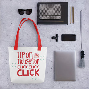 A tote bag lying on a surface with a laptop and office items next to it. The tote bag has the words "Up on the housetop, click, click, click" in red with three blue stars surroudning the words. 