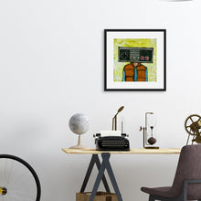 Load image into Gallery viewer, A black frame hanging above a desk featuring artwork with the original Nintendo controller as a &quot;head&quot; on a person wearing an orange puffer vest. 