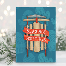 Load image into Gallery viewer, A Christmas card standing up with with pine leaves in the background with a touch of snow. The card has a blue background with lighter blue winter mittens in a pattern. On top of the mittens is an illustrated vintage sled with red ribbon and the words &quot;season&#39;s greetings&quot; in white.