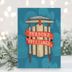 A Christmas card standing up with with pine leaves in the background with a touch of snow. The card has a blue background with lighter blue winter mittens in a pattern. On top of the mittens is an illustrated vintage sled with red ribbon and the words "season's greetings" in white.
