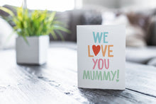 Load image into Gallery viewer, A greeting card featured on a black, wood coffee table. There’s a white planter in the background with a green plant. There’s also a gray sofa in the background with a white pillow. The card features the words “We Love You Mummy!” with the “O” in the word love featured as a heart.