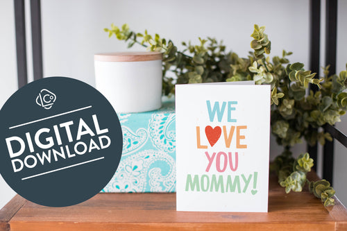 A greeting card is on a table top with a present in blue wrapping paper in the background. On top of the present is a candle and some greenery from a plant too. The card features the words “We Love You Mommy!” The words 