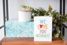 Load image into Gallery viewer, A greeting card is on a table top with a present in blue wrapping paper in the background. On top of the present is a candle and some greenery from a plant too. The card features the words “We Love You Mommy!” 