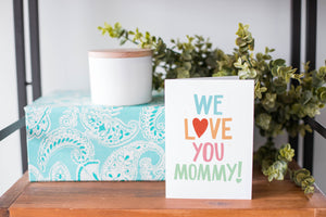 A greeting card is on a table top with a present in blue wrapping paper in the background. On top of the present is a candle and some greenery from a plant too. The card features the words “We Love You Mommy!” 