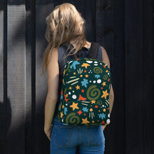 Load image into Gallery viewer, A woman standing in front of a black fence with her back to the camera. She has a backpack over both shoulders, resting on her back. The backpack is hunter green with a fun pattern of yellow stars, green swirls, blue &quot;splats&quot; and other fun whimsical shapes. The backpack straps are black. 