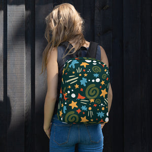 A woman standing in front of a black fence with her back to the camera. She has a backpack over both shoulders, resting on her back. The backpack is hunter green with a fun pattern of yellow stars, green swirls, blue "splats" and other fun whimsical shapes. The backpack straps are black. 