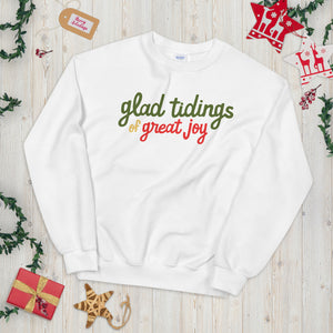 A white sweatshirt laying on a table with Christmas objects around it. The sweatshirt has the words "glad tidings of great joy" in red, green and yellow. 