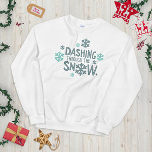 A white sweatshirt laying on a table with Christmas objects around it. The sweatshirt has the words "Dashing through the snow" in light and dark blue. There are snowflakes around the letters. 