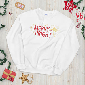 A white sweatshirt laying on a table with Christmas objects around it. The sweatshirt features the words Merry and Bright with illustrated Christmas stars around it. The words are in red and star illustrations are in yellow on the white fabric. 