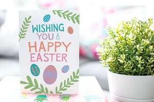 A photo of a card featured on a tabletop next to a white planter filled with a green plant. ​​The card features the words “Wishing you a happy Easter” with illustrated Easter eggs and palm leaves.