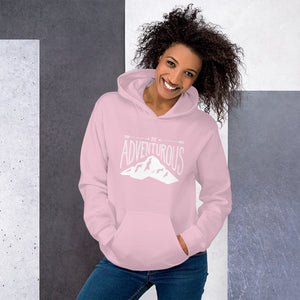 A woman wearing a light pink hoodie with lettering and illustration in white with the phrase “Be Adventurous” with arrows pointing to the word “be” and a mountain illustration underneath the word “adventure.”