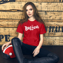 Load image into Gallery viewer, A woman wearing an red short sleeved t-shirt. The tee features lettering of the word &quot;Believe&quot; in white with the &quot;I&quot; of the word featured as an illustrated Christmas tree. 