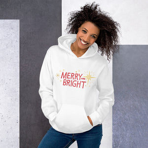 A woman wearing a white hoodie featuring hand drawn lettering in red with the words "Merry and Bright" with yellow illustrated stars around the words. 
