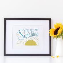 Load image into Gallery viewer, A white print in a black frame sits next to a white jug holding yellow sunflowers. The print reads &#39;You are my sunshine&#39; in blue lettering, over a yellow sun illustration