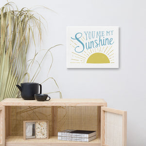 A white canvas hangs on a white wall over a wooden cane cabinet. The print reads 'You are my sunshine' in blue over a yellow sun illustration