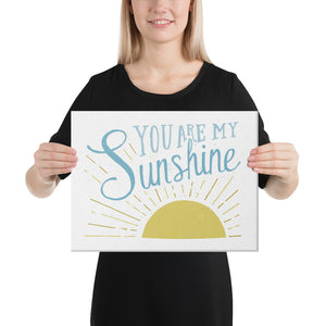 A woman is holding up a white canvas and smiling. The canvasreads 'You are my sunshine' in blue over a yellow sun illustration