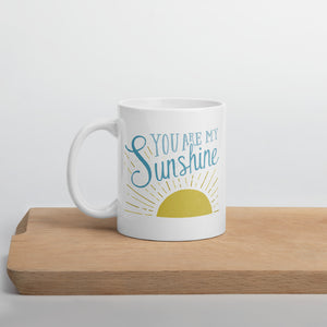 A white mug sits on a wooden shelf.  The mug reads 'You are my sunshine' in blue over a yellow sun illustration
