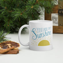 Load image into Gallery viewer, A white mug sits on a white table surrounded by evergreen branches and dried oranges.  The mug reads &#39;You are my sunshine&#39; in blue lettering over a yellow sun illustration