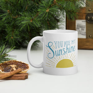 A white mug sits on a white table surrounded by evergreen branches and dried oranges.  The mug reads 'You are my sunshine' in blue lettering over a yellow sun illustration