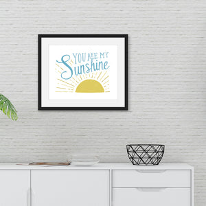 A large white print in a black frame hangs on a white painted brick wall, over a white sideboard. The print reads 'You are my sunshine' in blue lettering, over a yellow sun illustration.