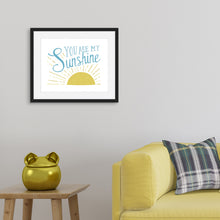 Load image into Gallery viewer, A white print in a black frame hangs on a pale grey living room wall, above a yellow sofa and wooden side table. The print reads &#39;You are my sunshine&#39; in blue lettering, over a yellow sun illustration.