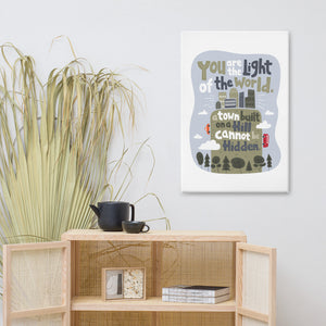 A canvas hanging on the wall above a shelving unit with a plant off to the side. The artwork is on a white background with lettering reading "You are the light of the world, a town built on a hill cannot be hidden." The words are a light gray background with an illustrated city.