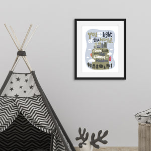 A picture frame with a black frame is featured on the wall of a playroom with a children's tent. The frame has illustrated artwork in it. The artwork is on a white background with lettering reading "You are the light of the world, a town built on a hill cannot be hidden." The words are a light gray background with an illustrated city.