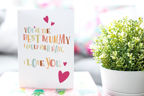 A greeting card is featured on a desktop with a green plant in the background. The card features illustrated lettering reading “You’re the best mummy I could ever have. I love you” with hearts surrounding the words. 